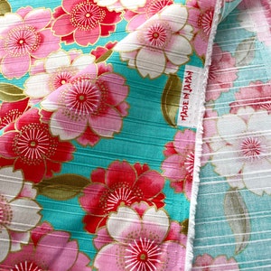 May include: A turquoise fabric with a pink and red floral pattern. The fabric has a ribbed texture and the flowers have gold accents. The fabric has a label that says "Made in Japan".