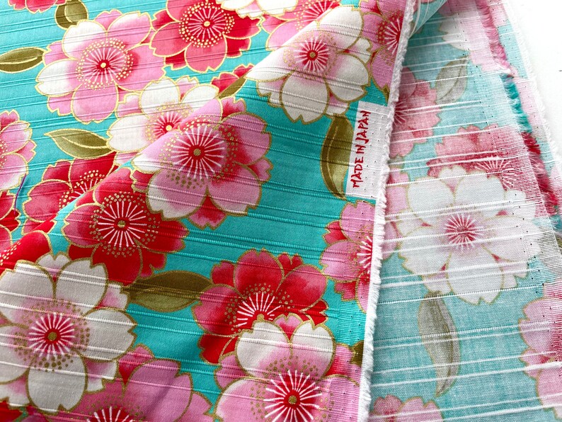 May include: A turquoise fabric with a pink and red floral pattern. The fabric has a ribbed texture and the flowers have gold accents. The fabric has a label that says "Made in Japan".