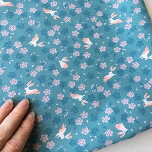 Peut inclure: Tissu bleu canard avec un motif r&eacute;p&eacute;t&eacute; de lapins blancs aux accents roses et de petites fleurs roses. Le tissu a un aspect textur&eacute; et convient &agrave; divers projets de couture.