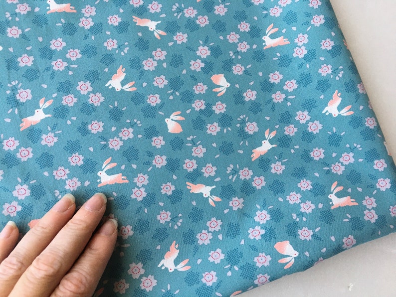 Peut inclure: Tissu bleu canard avec un motif r&eacute;p&eacute;t&eacute; de lapins blancs aux accents roses et de petites fleurs roses. Le tissu a un aspect textur&eacute; et convient &agrave; divers projets de couture.