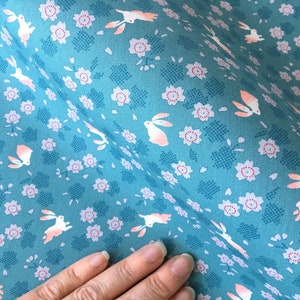 Peut inclure: Tissu bleu canard avec un motif r&eacute;p&eacute;t&eacute; de lapins blancs aux accents p&ecirc;che et de petites fleurs roses. Le tissu a un aspect textur&eacute;, id&eacute;al pour les projets de couture et d'artisanat.