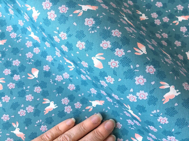 Peut inclure: Tissu bleu canard avec un motif r&eacute;p&eacute;t&eacute; de lapins blancs aux accents p&ecirc;che et de petites fleurs roses. Le tissu a un aspect textur&eacute;, id&eacute;al pour les projets de couture et d'artisanat.