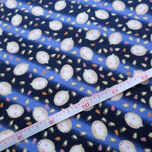 May include: Close-up of a fabric with a repeating pattern of white, oval-shaped figures with pink accents, scattered on a blue and navy striped background. A white measuring tape with red markings is laid across the fabric, showing measurements in inches.