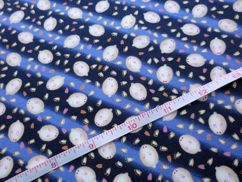 May include: Close-up of a fabric with a repeating pattern of white, oval-shaped figures with pink accents, scattered on a blue and navy striped background. A white measuring tape with red markings is laid across the fabric, showing measurements in inches.