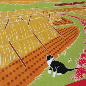 May include: A black and white cat sits in a field of red spider lilies. The cat is looking to the right. In the background, there are rows of hay bales and a train track.