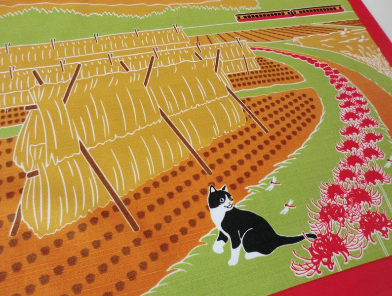 May include: A black and white cat sits in a field of red spider lilies. The cat is looking to the right. In the background, there are rows of hay bales and a train track.