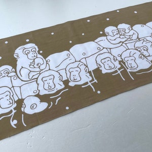 May include: A rectangular brown towel featuring a white illustration of several monkeys. The monkeys are depicted in a line, with some holding babies. White dots are scattered around the monkeys.