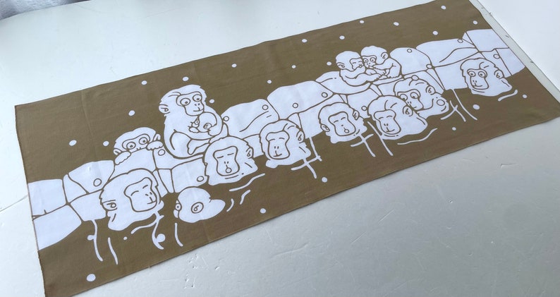 May include: A rectangular brown towel featuring a white illustration of several monkeys. The monkeys are depicted in a line, with some holding babies. White dots are scattered around the monkeys.