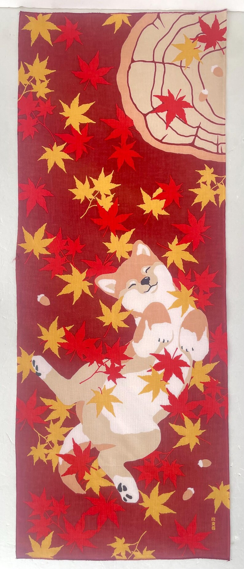 May include: A rectangular fabric wall hanging featuring a tan and white Shiba Inu dog lying on its back amidst a field of red and yellow maple leaves. The design includes a stylized tree stump in the upper corner and small acorns scattered throughout.