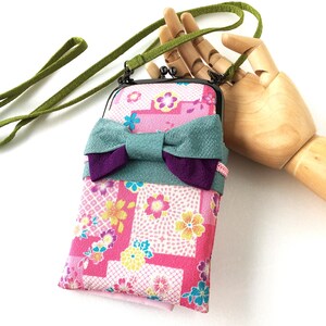 May include: A rectangular crossbody pouch featuring a pink fabric with a multi-colored floral pattern, accented by a large teal and purple fabric bow. The pouch has a black metal kiss-lock frame and a long, thin green fabric shoulder strap.