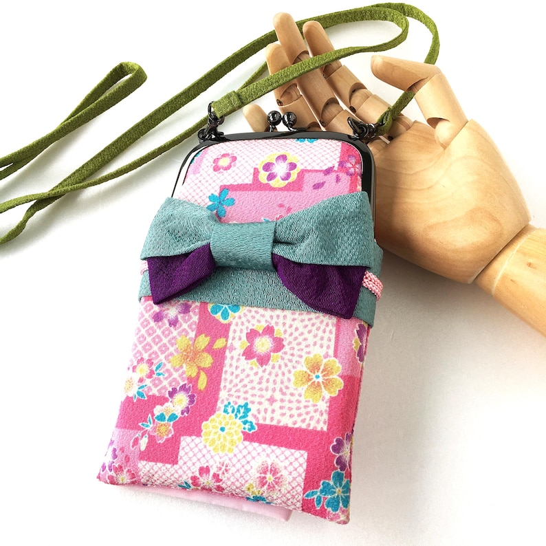 May include: A rectangular crossbody pouch featuring a pink fabric with a multi-colored floral pattern, accented by a large teal and purple fabric bow. The pouch has a black metal kiss-lock frame and a long, thin green fabric shoulder strap.