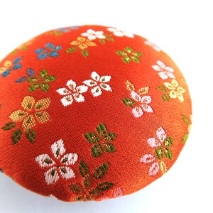 May include: A round, fabric-covered pincushion featuring a vibrant orange base color. The surface is decorated with an embroidered floral pattern consisting of small white, pink, blue, and green blossoms and leaves scattered across the fabric.