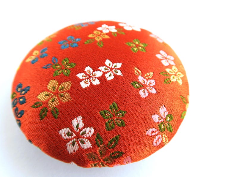 May include: A round, fabric-covered pincushion featuring a vibrant orange base color. The surface is decorated with an embroidered floral pattern consisting of small white, pink, blue, and green blossoms and leaves scattered across the fabric.