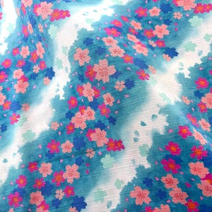 May include: A fabric featuring a vibrant teal and white watercolor-style background patterned with numerous small pink and dark blue cherry blossom flowers. The material has a visible horizontal texture throughout the entire surface.