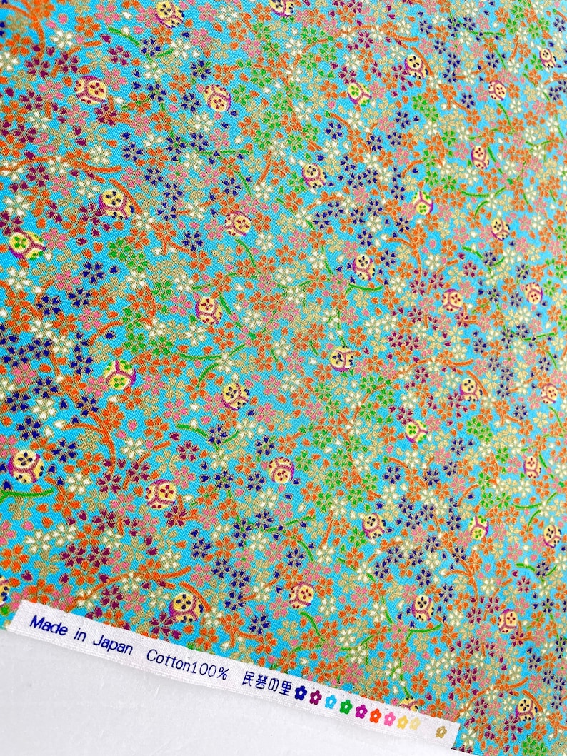 Puede incluir: Una tela azul brillante con un patr&oacute;n denso de peque&ntilde;as flores naranjas, blancas y azul oscuro, enredaderas verdes y motivos circulares. Un borde blanco en la parte inferior muestra el texto Made in Japan Cotton100% y caracteres japoneses.