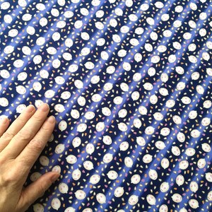 May include: A blue fabric with a repeating pattern of white, cartoon-like faces with small pink accents and scattered orange and pink shapes. The design is arranged in diagonal stripes.