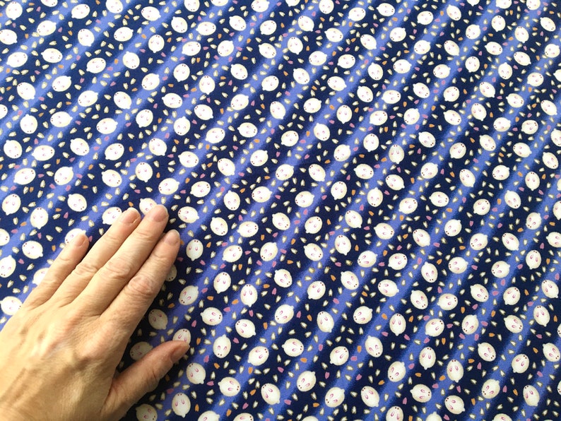 May include: A blue fabric with a repeating pattern of white, cartoon-like faces with small pink accents and scattered orange and pink shapes. The design is arranged in diagonal stripes.