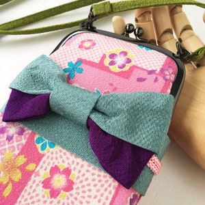 May include: A rectangular crossbody pouch featuring a pink fabric with a multi-colored floral pattern, accented by a large teal and purple fabric bow. The pouch has a black metal kiss-lock frame and a long, thin green fabric shoulder strap.