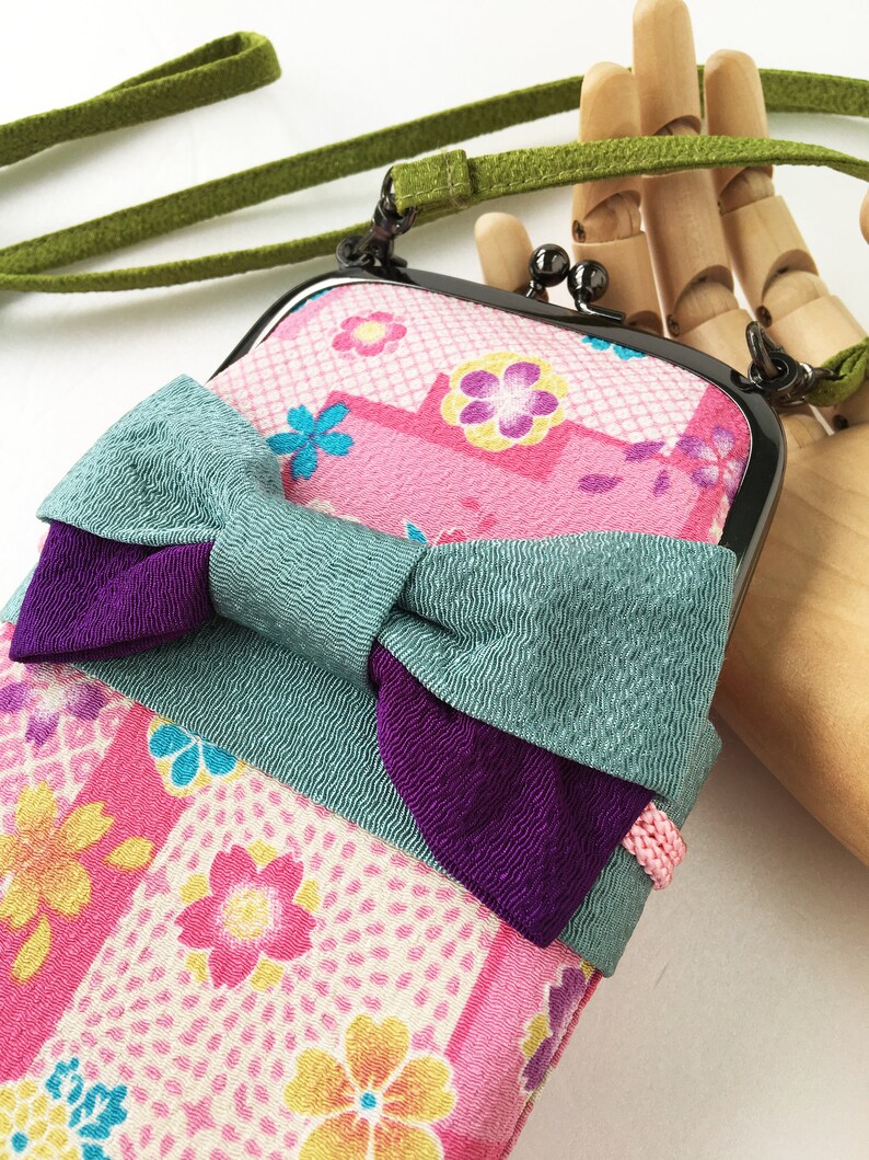 May include: A rectangular crossbody pouch featuring a pink fabric with a multi-colored floral pattern, accented by a large teal and purple fabric bow. The pouch has a black metal kiss-lock frame and a long, thin green fabric shoulder strap.