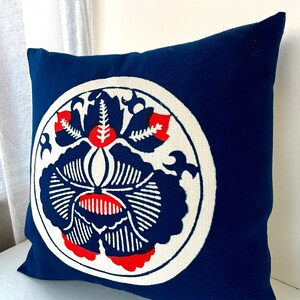 Op de afbeelding: Een blauw kussen met een witte cirkel en een rood en wit bloemenontwerp. Het kussen is gemaakt van een zachte, textuurrijke stof.