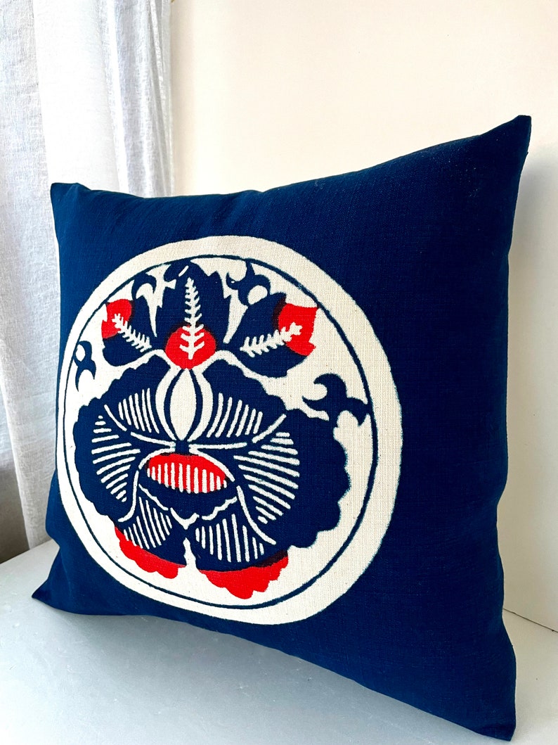 Op de afbeelding: Een blauw kussen met een witte cirkel en een rood en wit bloemenontwerp. Het kussen is gemaakt van een zachte, textuurrijke stof.