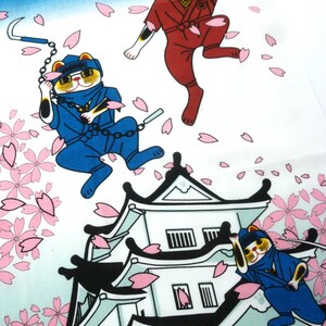 May include: A fabric print featuring three calico cats dressed as ninjas in red and blue uniforms. The cats are depicted jumping and climbing around a white Japanese castle, surrounded by falling pink cherry blossom petals against a blue sky.