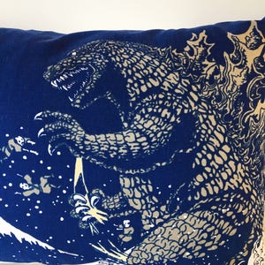 May include: Blue pillow with a white print of a Godzilla-like monster with a textured skin pattern. The monster is standing on a wave with a white background.