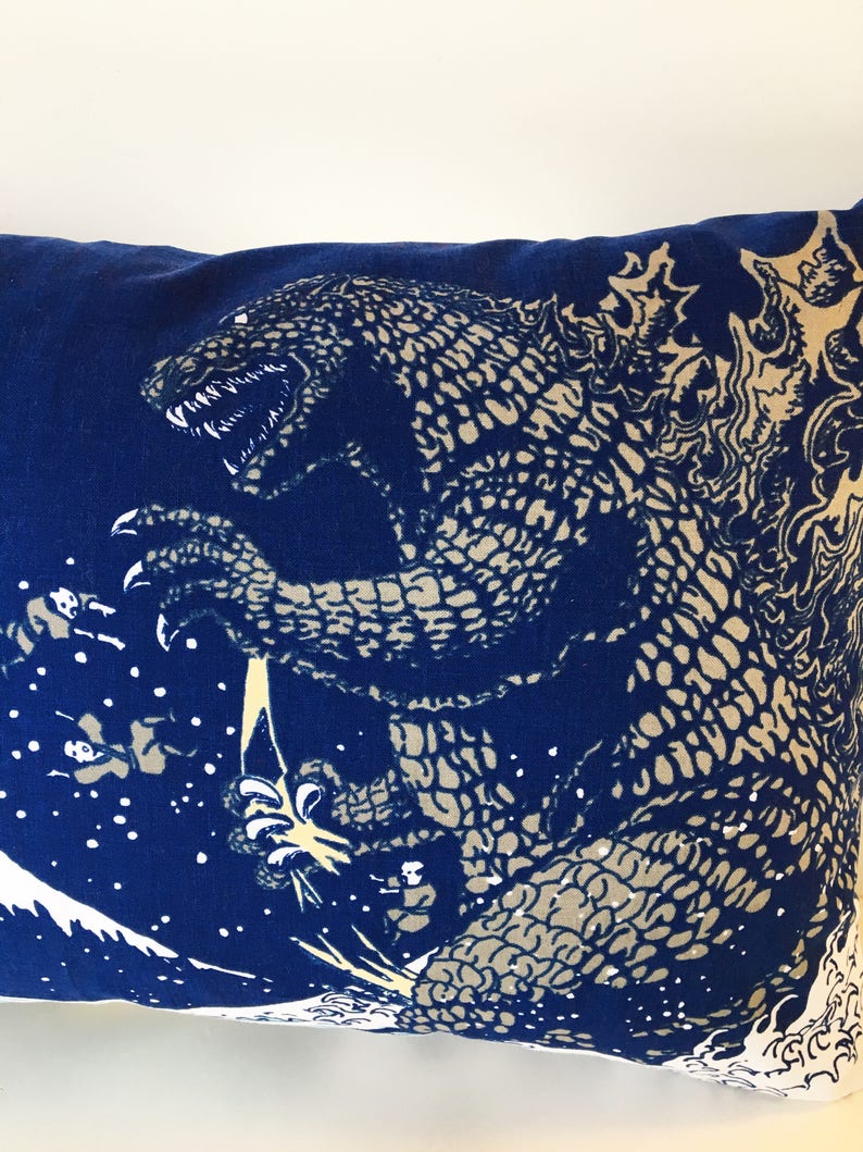 May include: Blue pillow with a white print of a Godzilla-like monster with a textured skin pattern. The monster is standing on a wave with a white background.