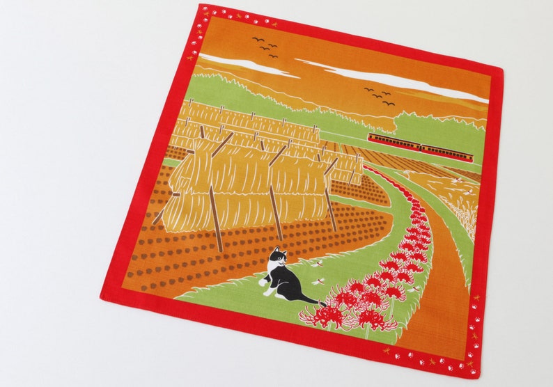 May include: A red fabric square with a colorful illustration of a rural landscape. The scene includes a field of hay bales, a winding path lined with red flowers, a train, and a black and white cat. The background features a sunset over a green hillside.