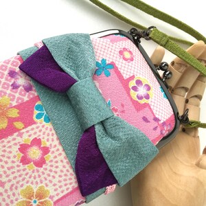 May include: A rectangular pouch with a dark metal kiss-lock clasp and a green fabric strap. The exterior features a pink fabric with a multi-colored floral pattern and a large, layered bow in textured teal and deep purple fabric across the front.