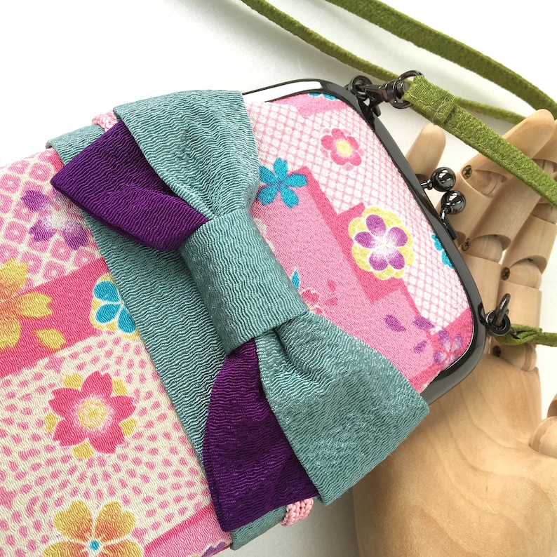 May include: A rectangular pouch with a dark metal kiss-lock clasp and a green fabric strap. The exterior features a pink fabric with a multi-colored floral pattern and a large, layered bow in textured teal and deep purple fabric across the front.