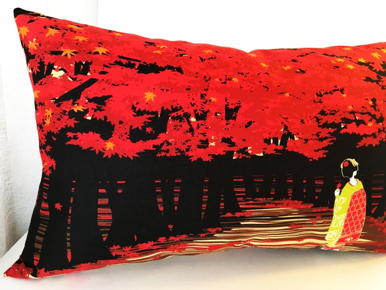 May include: A black and red pillow with a Japanese-inspired design featuring a woman in a yellow and red kimono standing in front of a red maple tree. The background is a black and brown striped pattern.