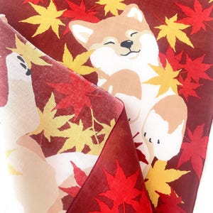 May include: A fabric textile featuring a repeating pattern of a smiling Shiba Inu dog surrounded by vibrant red and yellow maple leaves against a deep maroon background. The material is folded over to reveal the same graphic print on the reverse side.