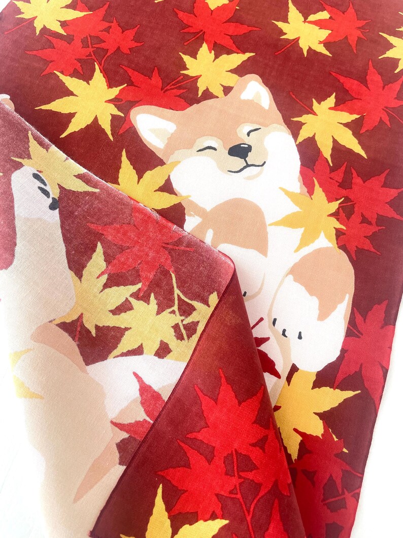 May include: A fabric textile featuring a repeating pattern of a smiling Shiba Inu dog surrounded by vibrant red and yellow maple leaves against a deep maroon background. The material is folded over to reveal the same graphic print on the reverse side.