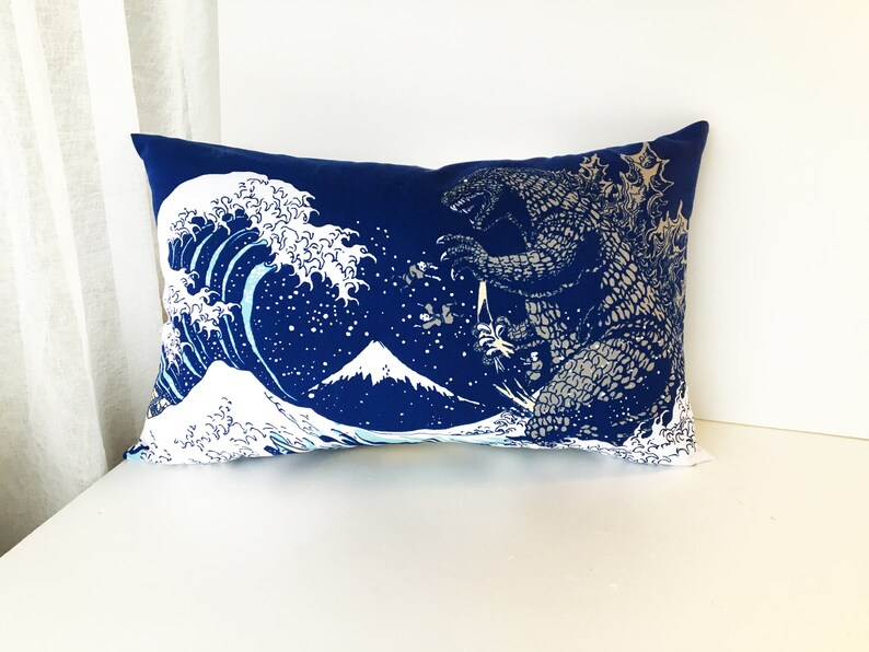 May include: A blue cushion featuring a white graphic of a wave, a mountain, and a Godzilla-like monster. The design is reminiscent of the famous Japanese woodblock print "The Great Wave off Kanagawa".