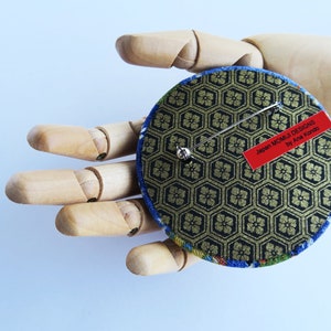 May include: A circular brooch featuring a black and gold hexagonal geometric pattern on the back, with a silver metal pin mechanism and a red rectangular label that reads Japan MOMIJI DESIGNS by Ana Kondo. The edges show blue and green fabric.