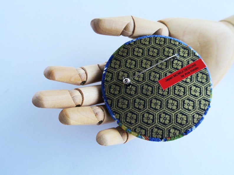 May include: A circular brooch featuring a black and gold hexagonal geometric pattern on the back, with a silver metal pin mechanism and a red rectangular label that reads Japan MOMIJI DESIGNS by Ana Kondo. The edges show blue and green fabric.