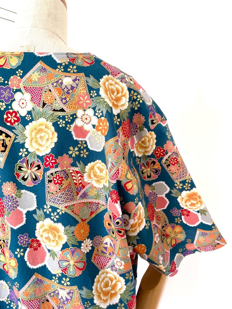 Puede incluir: Una camisa de manga corta azul turquesa con un estampado floral colorido. El estampado presenta grandes flores amarillas, flores rosas y formas geom&eacute;tricas.