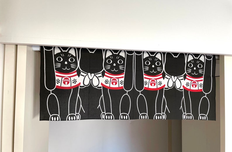 Puede incluir: Cortina Maneki Neko en blanco y negro. La cortina presenta un patr&oacute;n repetido de gatos negros estilizados con patas levantadas, collares rojos y detalles blancos. La cortina est&aacute; colgada en una puerta.