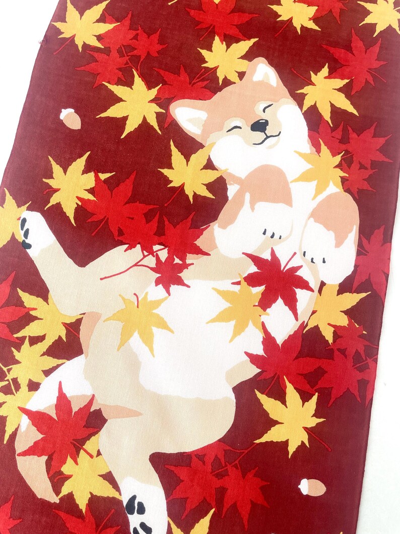 May include: A rectangular textile featuring a tan and white Shiba Inu dog lying on its back amidst a scattering of vibrant red and yellow maple leaves on a deep maroon background. The dog has a joyful expression with closed eyes and paws raised.