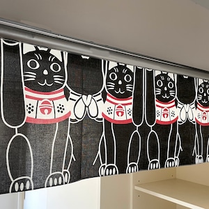 Puede incluir: Cortina japonesa Maneki Neko en blanco y negro. La cortina presenta un patr&oacute;n repetido de gatos estilizados con collares rojos y blancos. Los gatos levantan sus patas en un gesto de bienvenida. La cortina cuelga de una barra plateada.