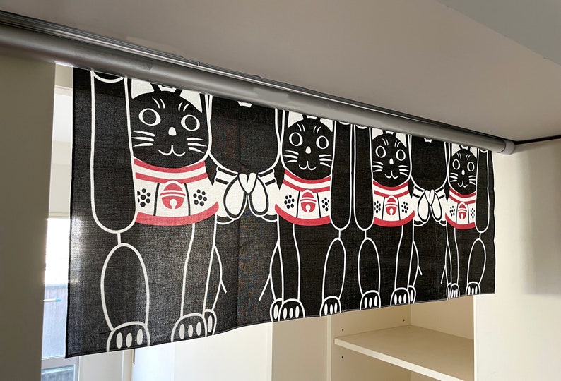 Puede incluir: Cortina japonesa Maneki Neko en blanco y negro. La cortina presenta un patr&oacute;n repetido de gatos estilizados con collares rojos y blancos. Los gatos levantan sus patas en un gesto de bienvenida. La cortina cuelga de una barra plateada.