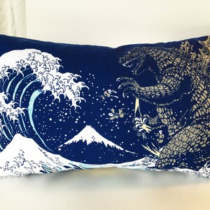 May include: A blue pillow with a white wave and a gray Godzilla monster on it. The wave is a stylized version of the Great Wave off Kanagawa, a famous Japanese woodblock print. The pillow is a decorative item for a home.