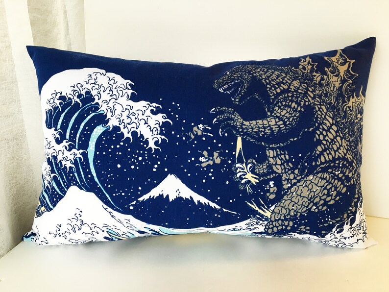 May include: A blue pillow with a white wave and a gray Godzilla monster on it. The wave is a stylized version of the Great Wave off Kanagawa, a famous Japanese woodblock print. The pillow is a decorative item for a home.