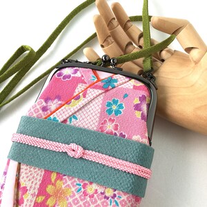 May include: A vertical crossbody pouch featuring a pink fabric with a multi-colored floral pattern, a dark metal kiss-lock clasp, a wide teal band across the center with a thin pink cord tied in a knot, and a long green suede-like shoulder strap.