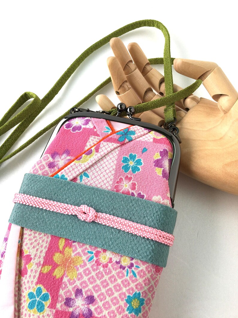 May include: A vertical crossbody pouch featuring a pink fabric with a multi-colored floral pattern, a dark metal kiss-lock clasp, a wide teal band across the center with a thin pink cord tied in a knot, and a long green suede-like shoulder strap.