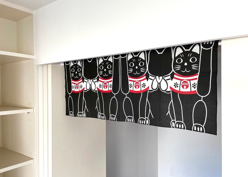 Puede incluir: Cortina japonesa Maneki Neko en blanco y negro. La cortina presenta un patr&oacute;n repetido de gatos negros estilizados con patas levantadas, collares rojos y detalles blancos. La cortina cuelga en una puerta.