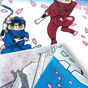 May include: A fabric featuring a pattern of cartoon cats dressed as ninjas in blue and maroon outfits, wielding weapons amidst falling pink cherry blossom petals against a white and blue gradient background, with a folded corner showing the reverse side.