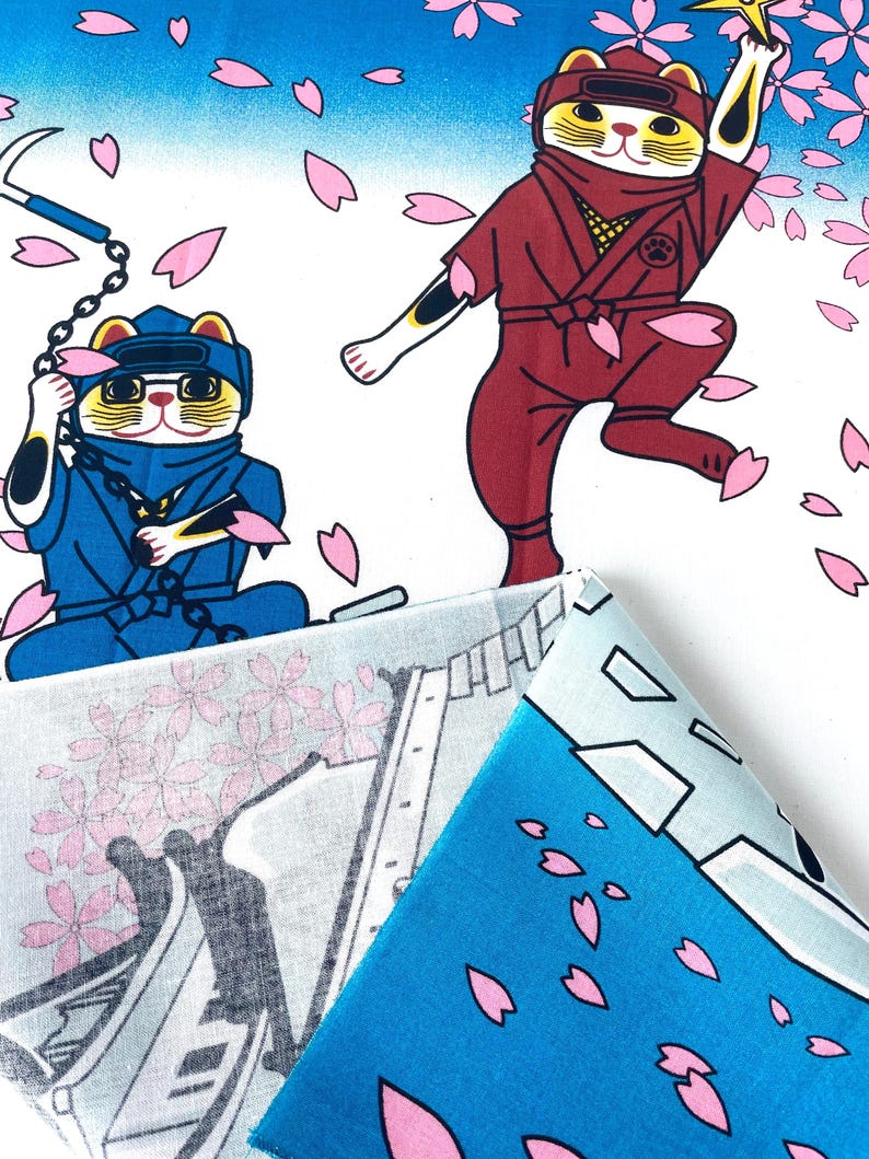 May include: A fabric featuring a pattern of cartoon cats dressed as ninjas in blue and maroon outfits, wielding weapons amidst falling pink cherry blossom petals against a white and blue gradient background, with a folded corner showing the reverse side.