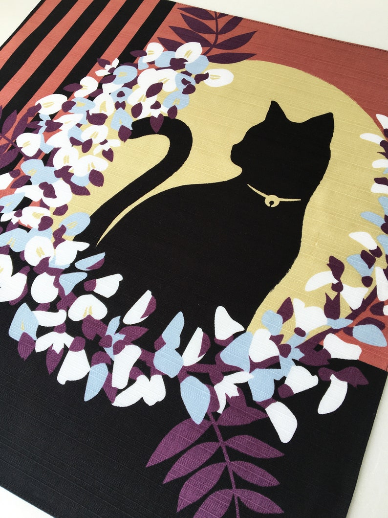 以下が含まれることがあります： 黒い猫のシルエットが、金色の首輪をつけて、白と青の花の輪の中に座っています。背景は、ピンクと黒のストライプ模様になっています。